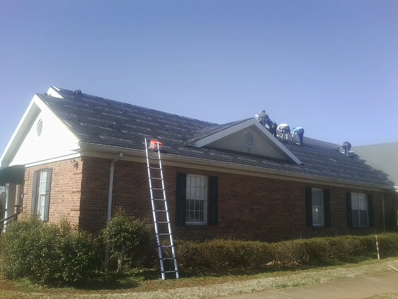 Crew tearing shingles off a brick ranch roof with a ladder set — Metal Roof Repair work by our crew in Cross Anchor