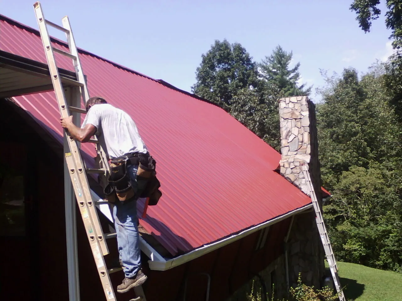 Roofer on a ladder beside a red metal roof and stone chimney — Soffit & Fascia Repair by Roofing Brothers in Cross Anchor